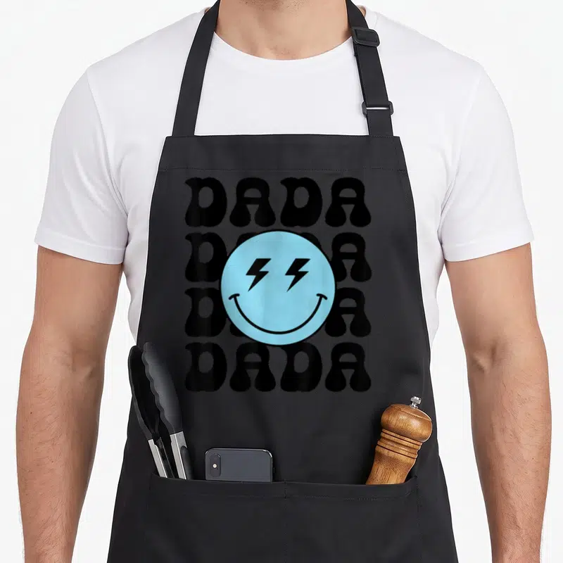 Family custom face apron - Light blue smiley face with black lightning eyes over four stacked DADA words in black — shown on a black cotton canvas apron against a white background