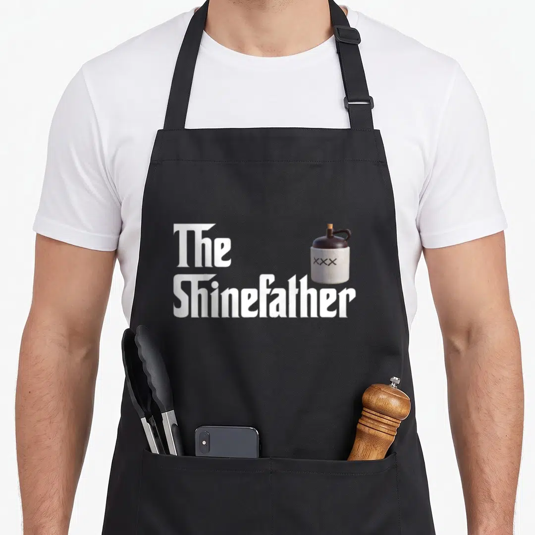 Shine father apron - White stacked text reading The Shinefather next to a brown and white moonshine jug with XXX — shown on a black cotton canvas apron against a white background