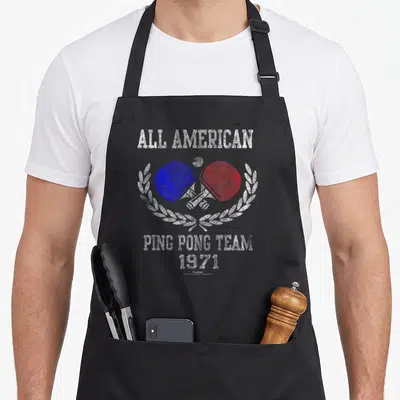 Forrest pong quotes apron - Crossed blue and red ping pong paddles in white laurel wreath with ALL AMERICAN text — shown on a black cotton canvas apron against a white background