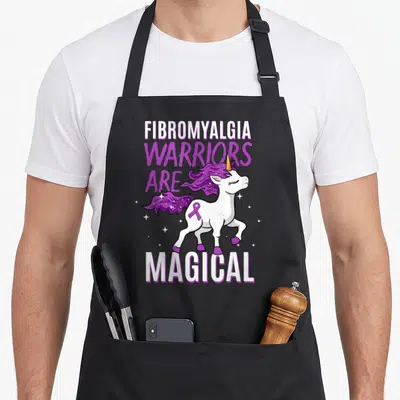 Fibromyalgia unicorn apron - Cartoon white unicorn with purple mane, purple ribbon, and stacked text reading warriors — shown on a black cotton canvas apron against a white background