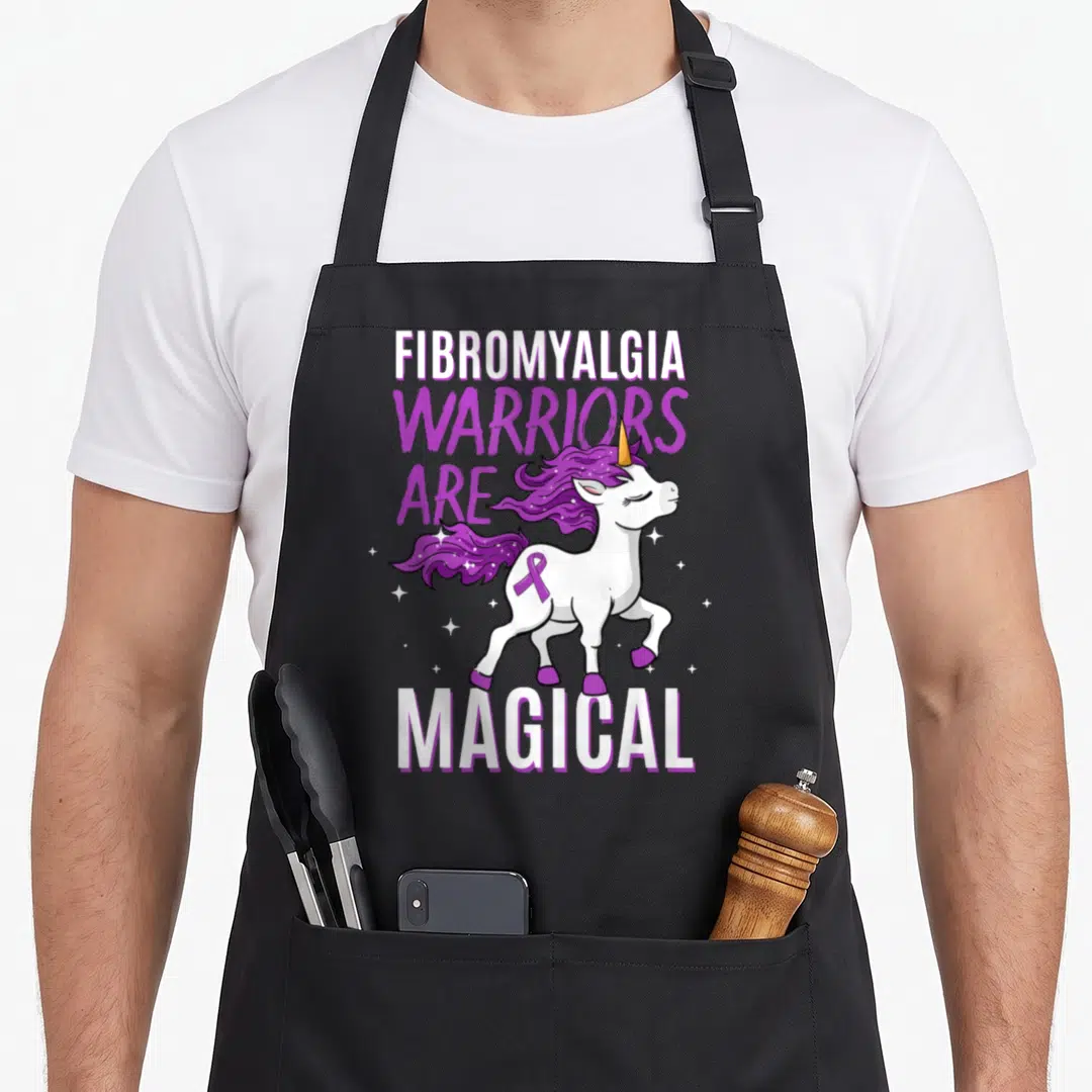 Fibromyalgia unicorn apron - Cartoon white unicorn with purple mane, purple ribbon, and stacked text reading warriors — shown on a black cotton canvas apron against a white background