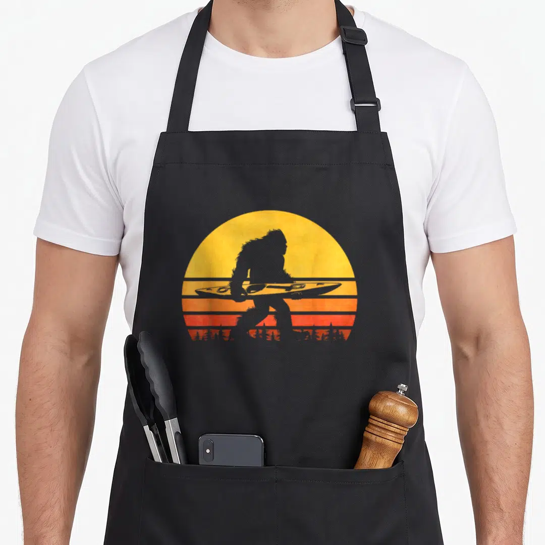 Kayak bigfoot apron - Bigfoot silhouette holding a kayak in front of a yellow and orange striped retro sun — shown on a black cotton canvas apron against a white background