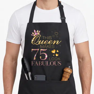 Queen fabulous birthday apron - Gold and pink rhinestone text reading This Queen Makes 75 Look Fabulous with crown — shown on a black cotton canvas apron against a white background