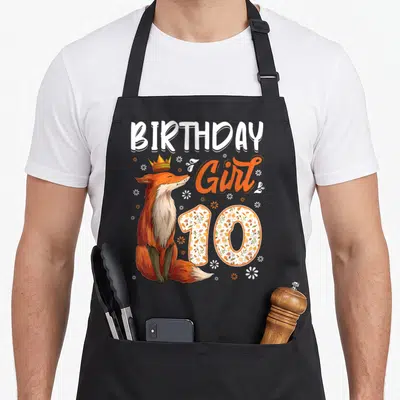 Animal fox print apron - Cartoon fox with gold crown next to BIRTHDAY Girl 10 text and scattered white flowers — shown on a black cotton canvas apron against a white background