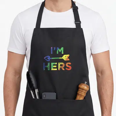 Lesbian couple apron - I'M HERS rainbow arrow text with heart tip in blue green yellow orange red — shown on a black cotton canvas apron against a white background