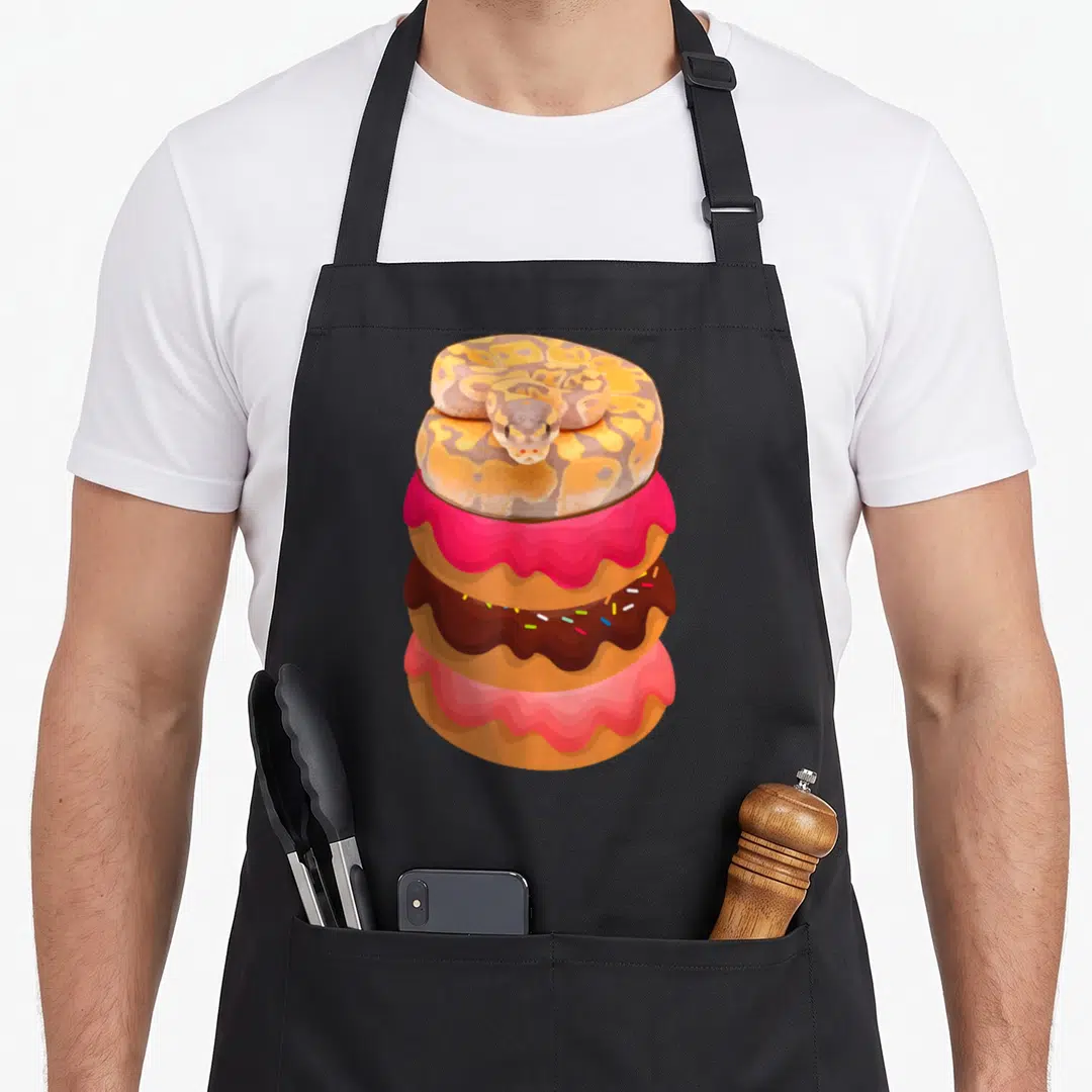 Snake donut apron - Cartoon snake coiled on three stacked doughnuts with pink and brown icing — shown on a black cotton canvas apron against a white background