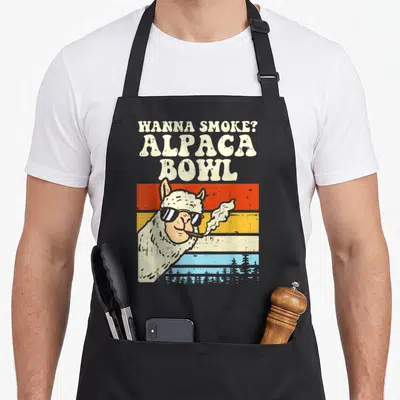 Smoke alpaca apron - Retro alpaca wearing sunglasses smoking in front of orange, yellow, and blue horizontal stripes — shown on a black cotton canvas apron against a white background
