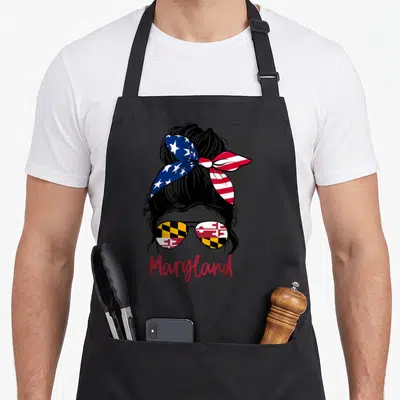 Maryland messy aprons - Cartoon woman with bun, US bandana, Maryland flag sunglasses, red Maryland text — shown on a black cotton canvas apron against a white background