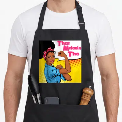 Melanin business apron - Cartoon woman flexing arm with That Melanin Tho text in pink speech bubble on yellow background — shown on a black cotton canvas apron against a white background