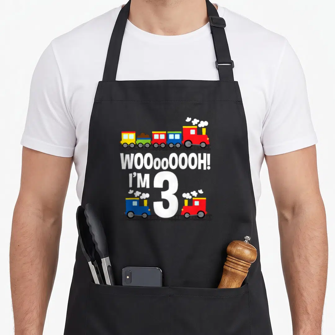 Woooooooh birthday apron - Colorful cartoon trains on tracks surrounding white text reading WOOoOOOOH I M 3 — shown on a black cotton canvas apron against a white background