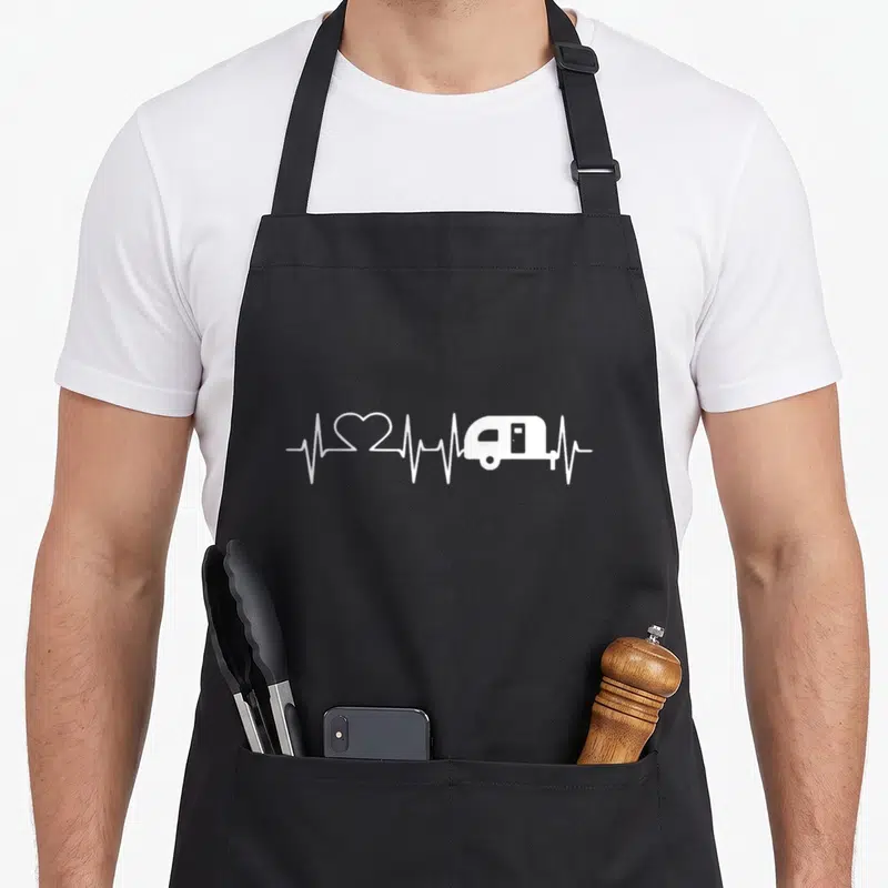 Heartbeat happy camper apron - White heartbeat line graphic merging a heart shape and camper trailer silhouette — shown on a black cotton canvas apron against a white background