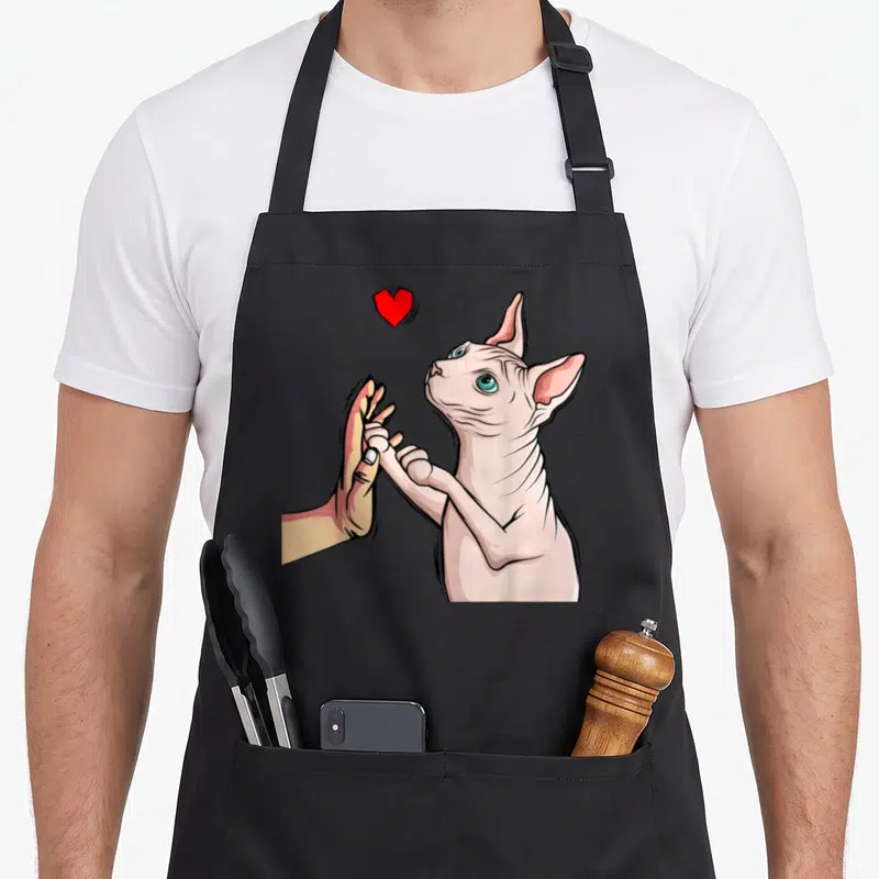 Sphinx cat apron - Pink sphynx cat high-fiving human hand with red heart cartoon — shown on a black cotton canvas apron against a white background