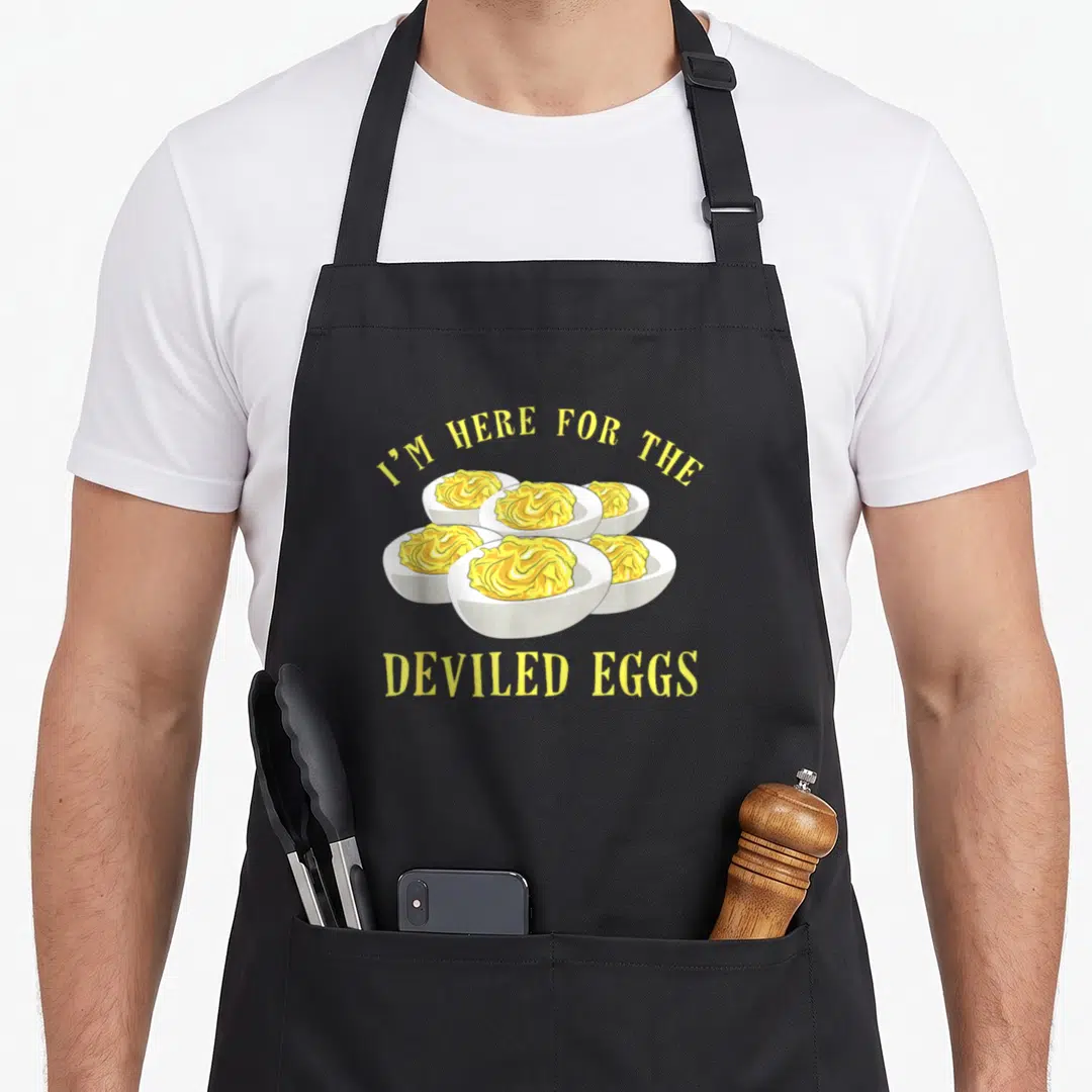 Food egg apron - Cartoon cluster of six deviled eggs with yellow filling and arched text — shown on a black cotton canvas apron against a white background