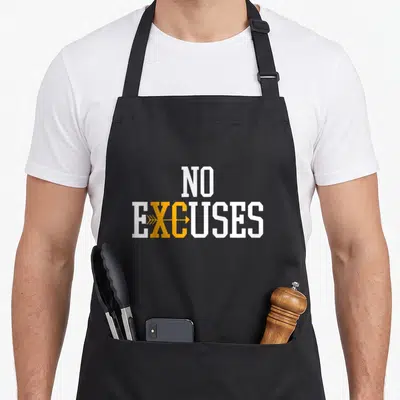 Excuse country apron - White NO EXCUSES text with orange arrow overlaying X and C letters — shown on a black cotton canvas apron against a white background