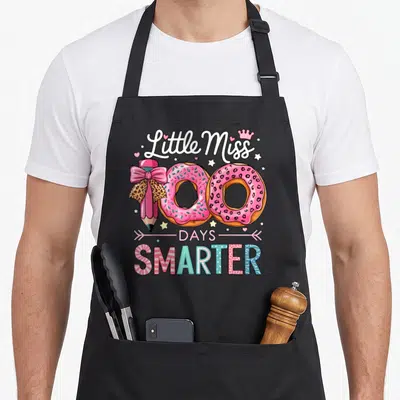 Miss love kindergarten apron - Little Miss 100 Days Smarter text with two pink donuts and pencil forming the number 100 — shown on a black cotton canvas apron against a white background