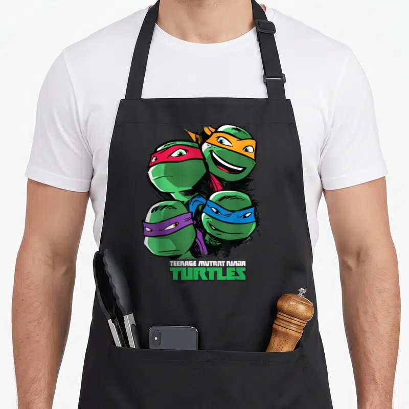 Character face teenage cooking aprons - Four cartoon turtle heads with red orange blue and purple masks above stacked — shown on a black cotton canvas apron against a white background