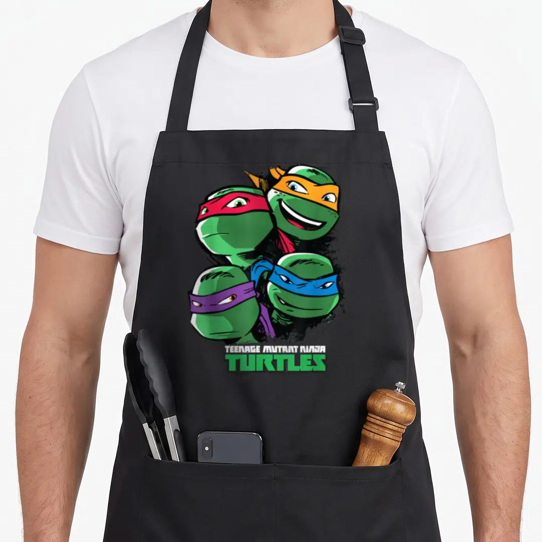 Character face teenage cooking aprons - Four cartoon turtle heads with red orange blue and purple masks above stacked — shown on a black cotton canvas apron against a white background
