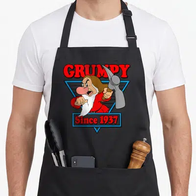 Triangle snow white apron - Grumpy character holding pickaxe in blue triangle with Since 1937 text — shown on a black cotton canvas apron against a white background