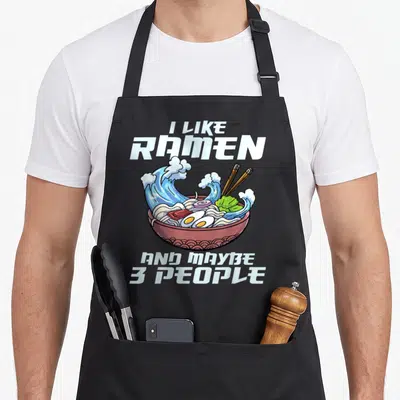 Japanese noodle anime apron - Cartoon ramen bowl with blue waves and text I like ramen and maybe 3 people — shown on a black cotton canvas apron against a white background