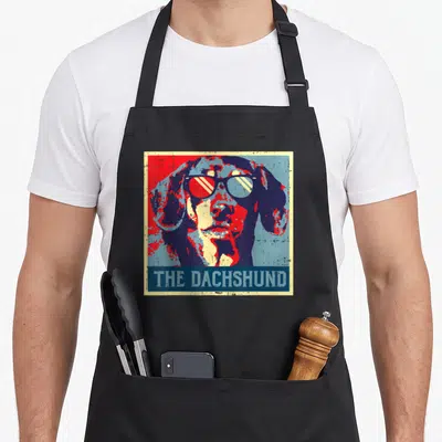Poster sausage apron - Retro dachshund portrait in red blue yellow with sunglasses and text THE DACHSHUND on blue banner — shown on a black cotton canvas apron against a white background