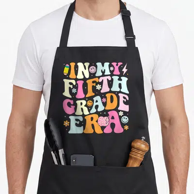 Grade era teacher apron - Stacked retro text IN MY FIFTH GRADE ERA with colorful pencils flowers and smiley faces — shown on a black cotton canvas apron against a white background
