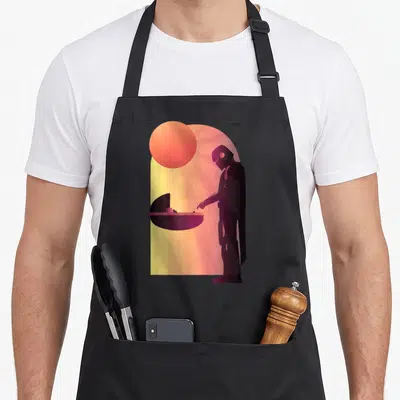 Epic consistency apron - Minimalist cloaked figure reaching toward a cradle under a large sun in purple orange — shown on a black cotton canvas apron against a white background