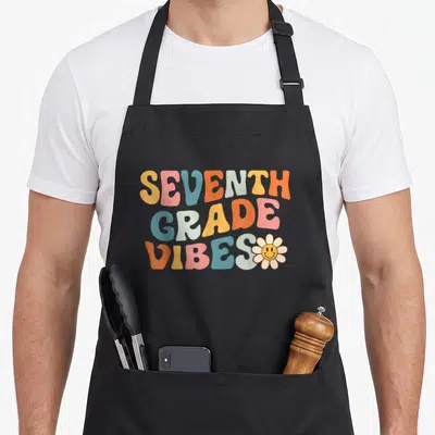 Seventh grade vibes apron - Retro text SEVENTH GRADE VIBES in orange pink blue beige with smiling flower — shown on a black cotton canvas apron against a white background