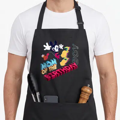 Funny star family aprons - Leaping Mickey Mouse above 3D text MOM OF THE BIRTHDAY BOY with rainbow streak — shown on a black cotton canvas apron against a white background