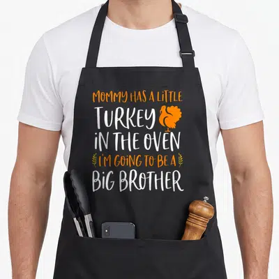 Brother pregnancy thanksgiving apron - MOMMY HAS A LITTLE TURKEY IN THE OVEN I'M GOING TO BE A BIG BROTHER text — shown on a black cotton canvas apron against a white background
