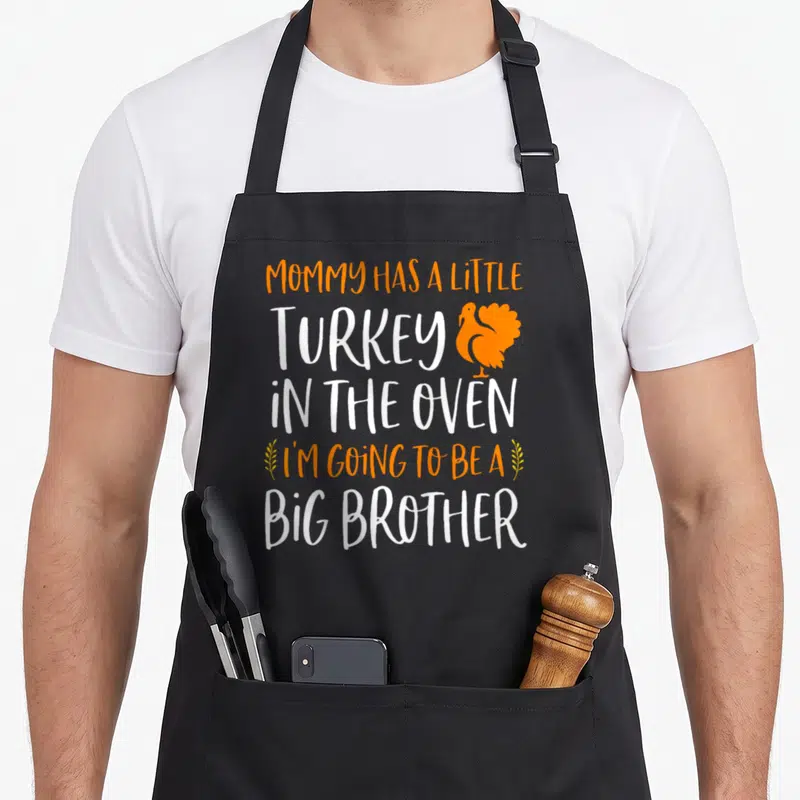 Brother pregnancy thanksgiving apron - MOMMY HAS A LITTLE TURKEY IN THE OVEN I'M GOING TO BE A BIG BROTHER text — shown on a black cotton canvas apron against a white background
