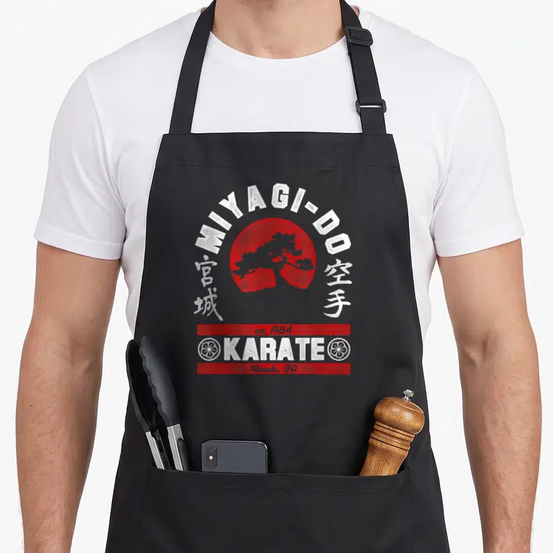 Arched tree apron - Miyagi-Do karate logo with red sun, bonsai tree, and Reseda CA text in retro red and white — shown on a black cotton canvas apron against a white background