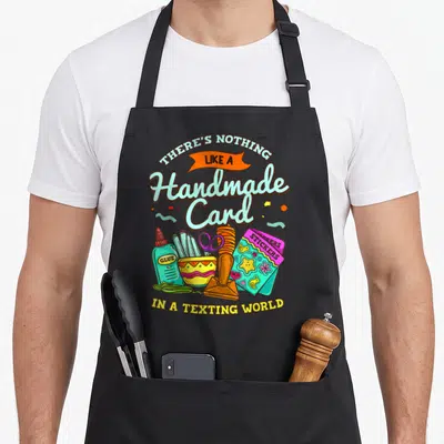 Card crafting apron - Cartoon glue bottle, purple scissors, orange stamp, sticker pack, and arched typography — shown on a black cotton canvas apron against a white background