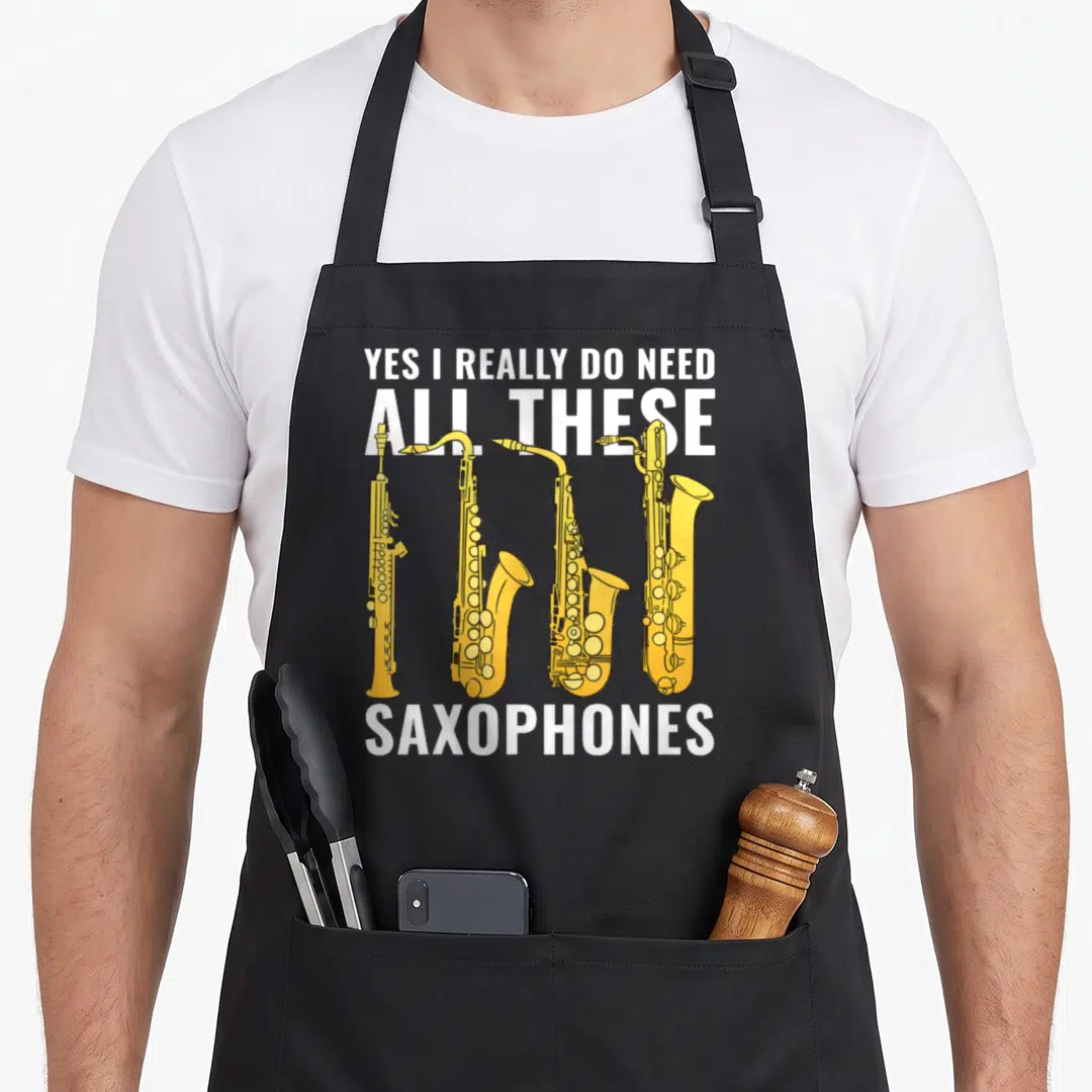These novelty apron - Four yellow cartoon saxophones with white text Yes I Really Do Need All These Saxophones — shown on a black cotton canvas apron against a white background
