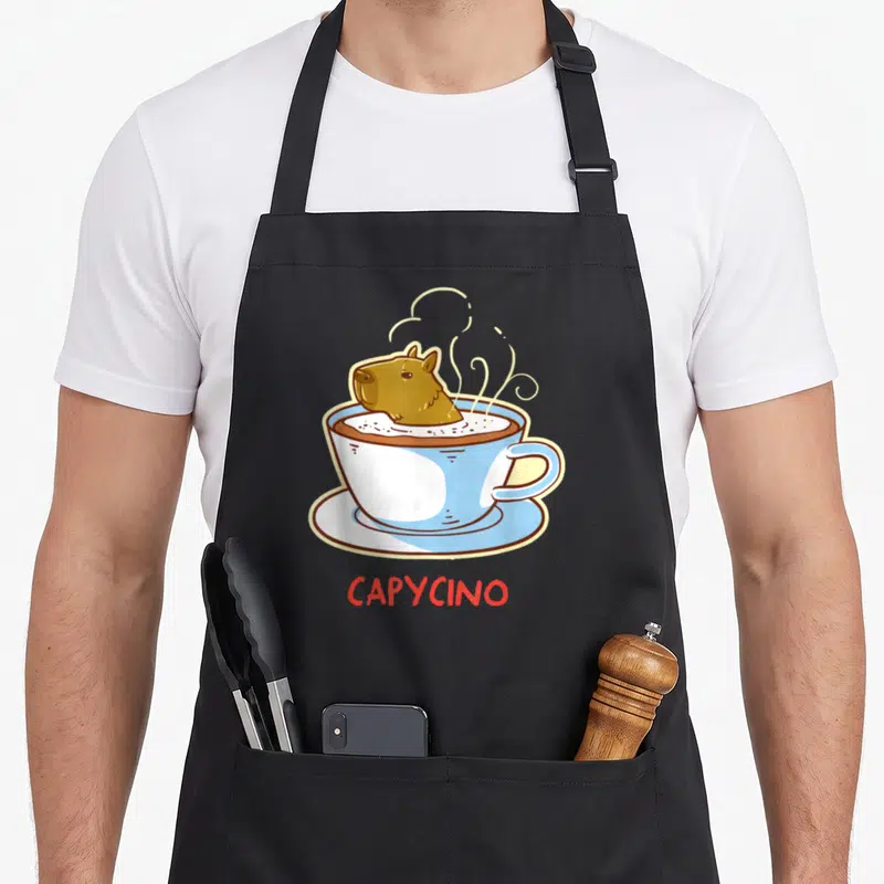 Capybara coffee apron - Cartoon brown capybara in blue cup with red CAPYCINO text below — shown on a black cotton canvas apron against a white background