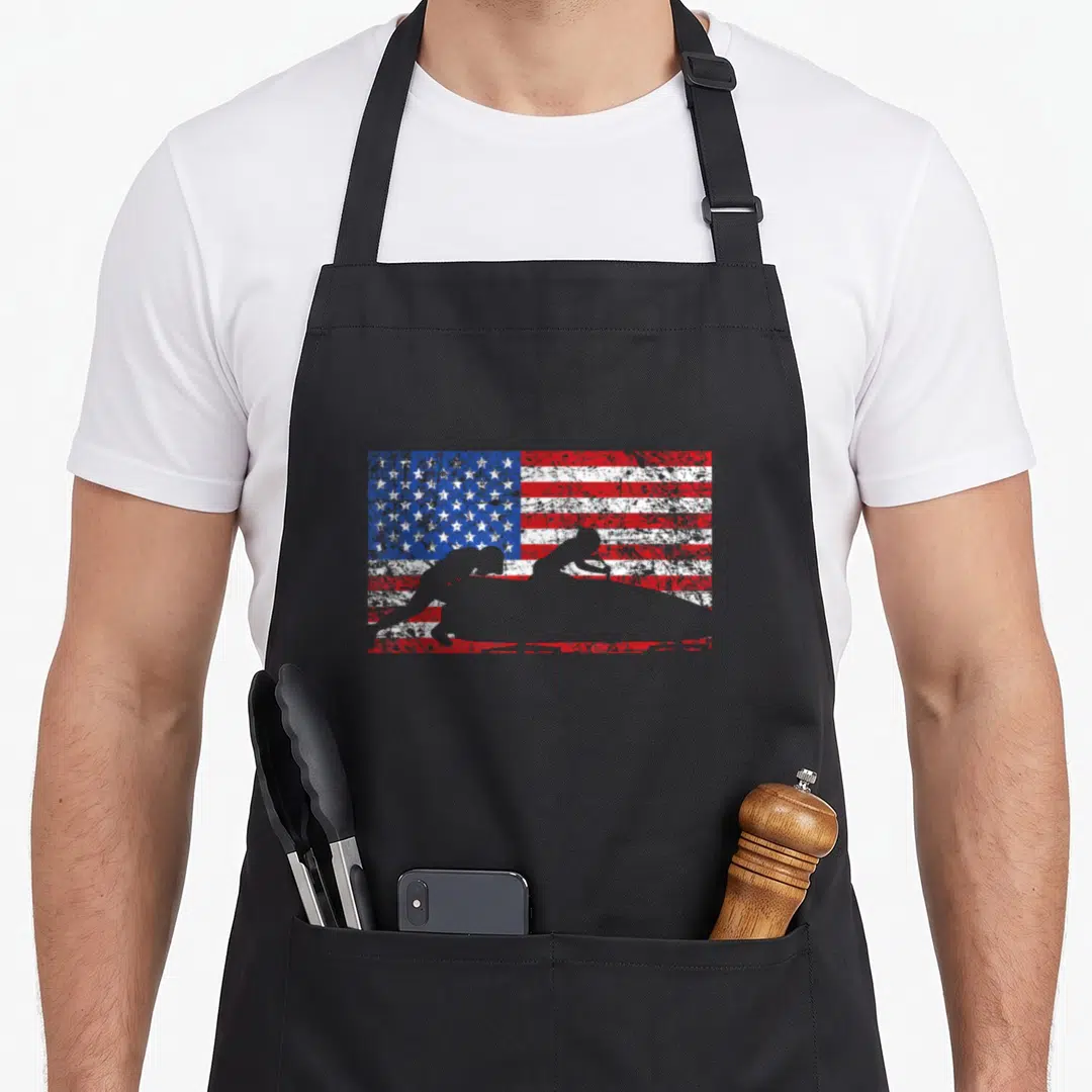 Bobsledding american flag apron - Two grey surfer silhouettes on a surfboard over a distressed American flag — shown on a black cotton canvas apron against a white background