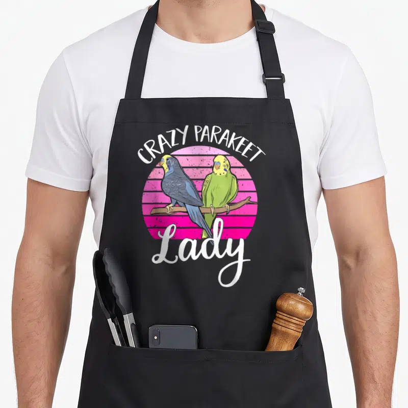 Parakeet pet apron - Two parakeets on a branch before a striped pink circle with white text reading CRAZY PARAKEET Lady — shown on a black cotton canvas apron against a white background