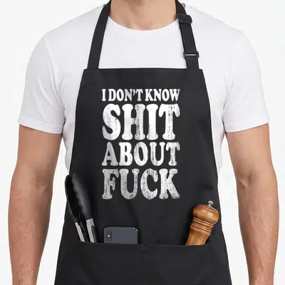 Profanity dump apron - White stacked typography reading I DON'T KNOW SHIT ABOUT FUCK on black fabric — shown on a black cotton canvas apron against a white background