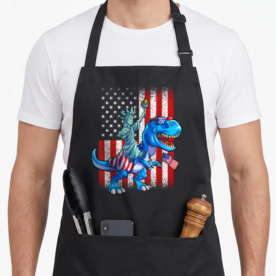 Dino 4th of july apron - Blue T-Rex with sunglasses and flag ridden by Statue of Liberty on distressed flag — shown on a black cotton canvas apron against a white background