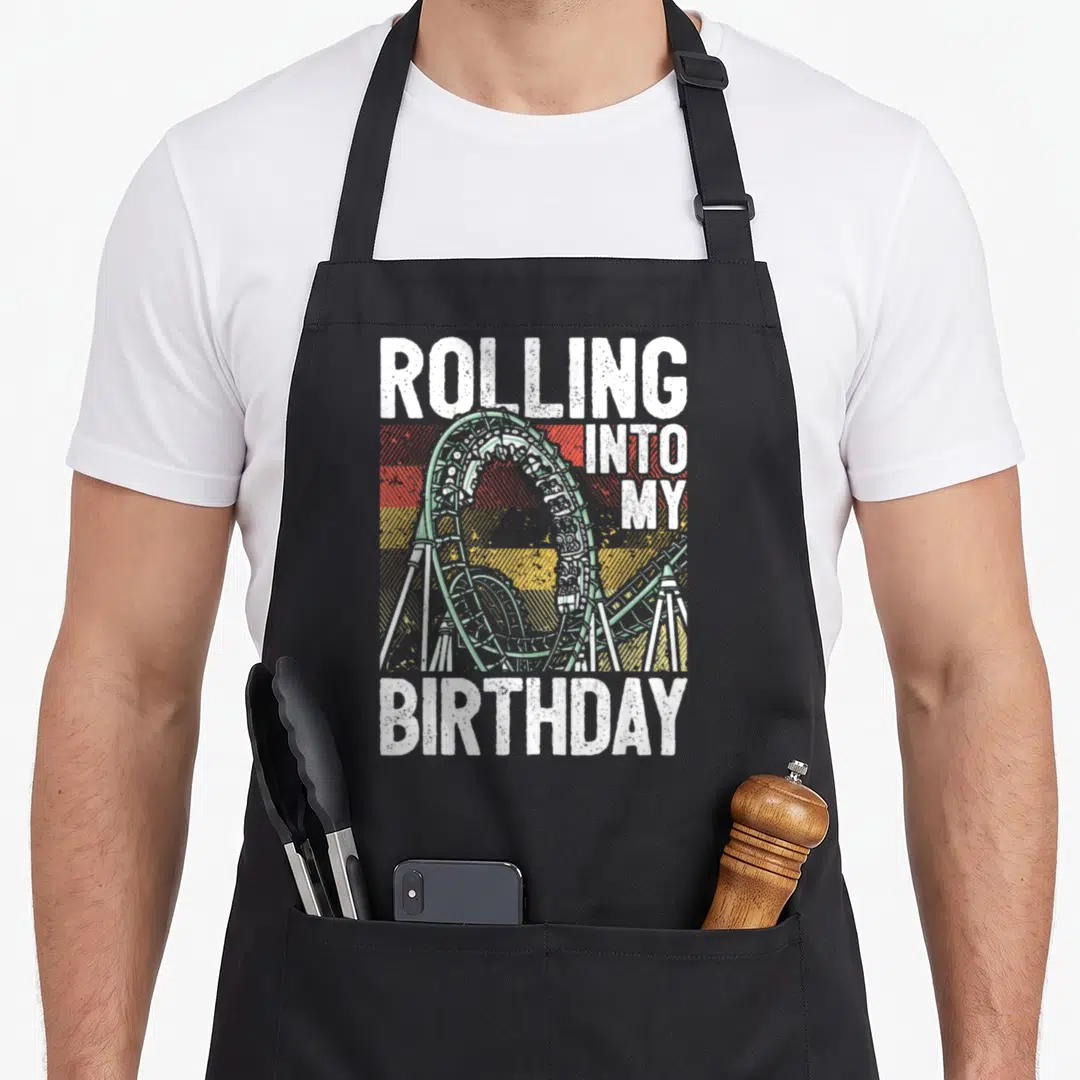 Park birthday apron - Green roller coaster loop over red orange yellow stripes with white distressed text — shown on a black cotton canvas apron against a white background