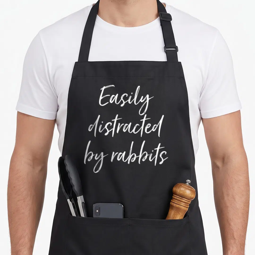 Easily rabbit apron - White stacked script text reading Easily distracted by rabbits — shown on a black cotton canvas apron against a white background