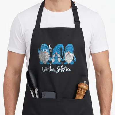 Gnome winter apron - Three blue gnomes holding hands under moon and snowflakes with Winter Solstice text — shown on a black cotton canvas apron against a white background