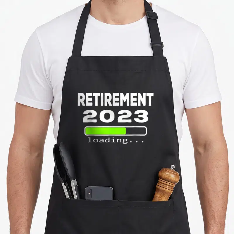 Loading retirement apron - White text reading RETIREMENT 2023 above a green loading bar and small loading label — shown on a black cotton canvas apron against a white background