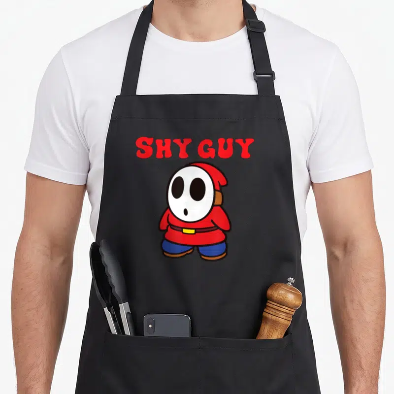 Bro simple apron - Red SHY GUY text above a white-masked cartoon character in red robe and blue pants — shown on a black cotton canvas apron against a white background