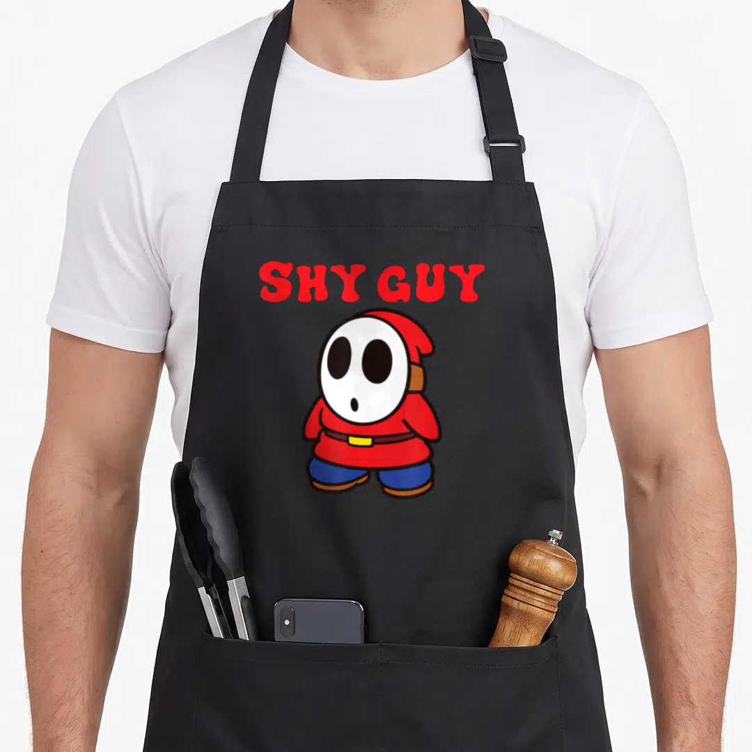 Bro simple apron - Red SHY GUY text above a white-masked cartoon character in red robe and blue pants — shown on a black cotton canvas apron against a white background