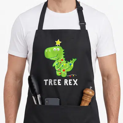Animal christmas tree apron - Green dinosaur wearing string lights and a yellow star on head above white text TREE REX — shown on a black cotton canvas apron against a white background