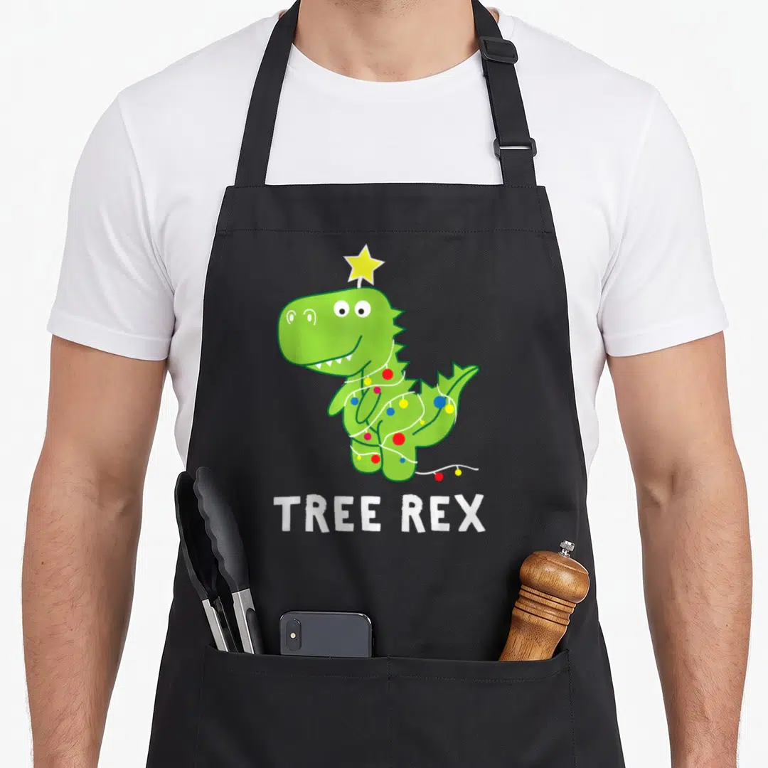 Animal christmas tree apron - Green dinosaur wearing string lights and a yellow star on head above white text TREE REX — shown on a black cotton canvas apron against a white background