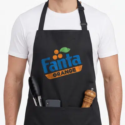 Coca orange soda apron - Distressed Fanta ORANGE logo with blue text, three orange circles, green leaf, and orange — shown on a black cotton canvas apron against a white background