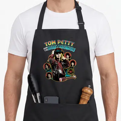 Tom caravan apron - Tom Petty tipping top hat surrounded by four circular band member portraits with arched retro text — shown on a black cotton canvas apron against a white background