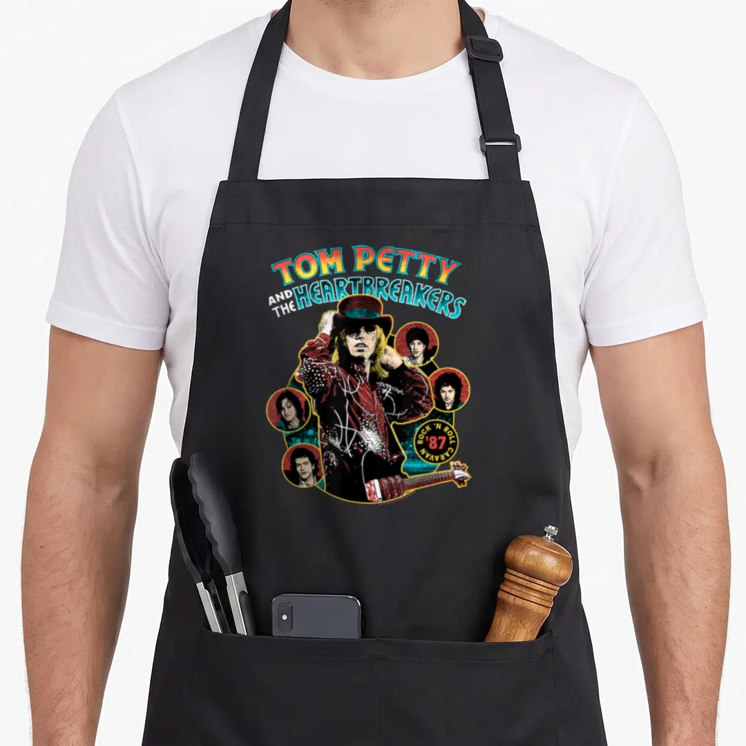 Tom caravan apron - Tom Petty tipping top hat surrounded by four circular band member portraits with arched retro text — shown on a black cotton canvas apron against a white background