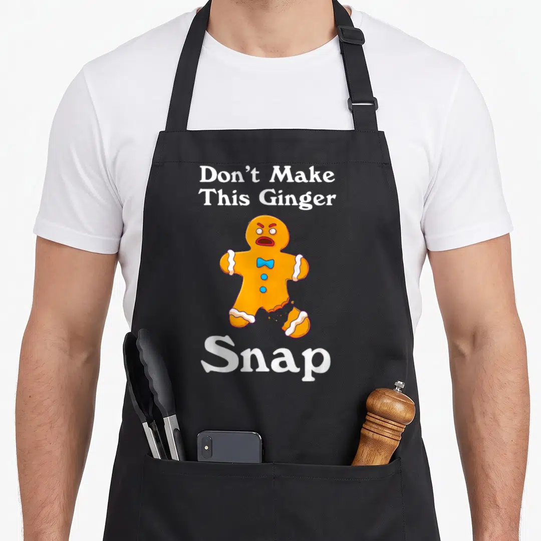 Snap christmas apron - Don't Make This Ginger Snap text above orange gingerbread man with broken leg and blue bowtie — shown on a black cotton canvas apron against a white background